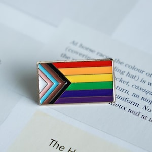 May include: A rectangular enamel pin with a gold border, displaying the Progress Pride flag design. The flag incorporates the rainbow stripes, plus a chevron of colours representing marginalized communities. The pin is on a white surface.