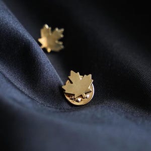 Könnte beinhalten: Zwei goldfarbene Ahornblatt-Anstecknadeln auf einem dunkelblauen Stoff. Die Anstecknadeln sind mit einem Ahornblatt-Design versehen und dienen wahrscheinlich als Accessoires. Der Stoff hat eine subtile Textur.