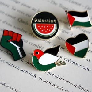May include: A collection of five enamel pins featuring the colors of the Palestinian flag. The set includes a round pin with a watermelon graphic and the text Palestine, a flag-shaped pin, a heart-shaped pin, a raised fist, and a bird.