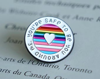Può includere: Spilla rotonda in smalto con bordo bianco e la scritta "YOU'RE SAFE TO BE YOU AROUND ME". La spilla presenta un design arcobaleno con un cuore bianco al centro, che simboleggia l'inclusione e il supporto.