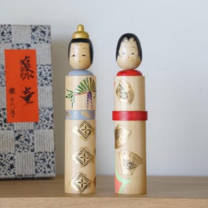 May include: Two Japanese Kokeshi dolls with painted faces and cylindrical bodies. One doll has a gold crown and green and purple painted details. The other doll has a red band and gold fan designs. A box with Japanese text is to the left.
