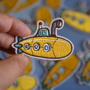 Peut inclure: Un écusson brodé représentant un sous-marin jaune avec un contour brun et trois hublots. Le sous-marin a un périscope bleu et blanc et une antenne rouge.