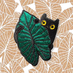Può includere: Toppa ricamata raffigurante un gatto nero che sbuca da dietro una grande foglia verde. Il gatto ha gli occhi gialli e la foglia ha cuciture dettagliate. La toppa è per abbigliamento o accessori.