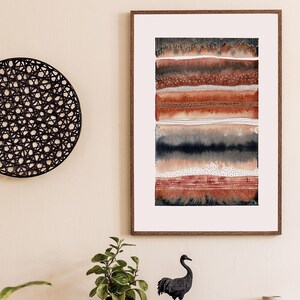May include: A framed abstract watercolor painting with brown, red, and black stripes. The painting is hanging on a beige wall above a wooden dresser with three drawers. A black ceramic vase with a plant and a black crane statue are on top of the dresser.