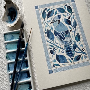 May include: A watercolor painting of a blue jay bird in a blue and white geometric frame. The bird is perched on a branch with leaves and berries. The painting is done in a folk art style.