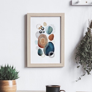 May include: Abstract art print in a light wood frame. The artwork features a collection of colorful, organic shapes in shades of blue, orange, and gray. The shapes are layered and textured, creating a visually interesting composition. The print is displayed on a white wall.