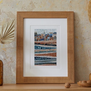 May include: A framed print of a watercolor painting depicting a stylized cityscape with blue and brown hues. The cityscape is set against a background of layered, textured stripes in shades of blue, brown, and gold.
