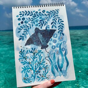 May include: Watercolor painting of a blue stingray with white spots, surrounded by blue and white seaweed and bubbles. The painting is on a white sheet of paper with a spiral binding.