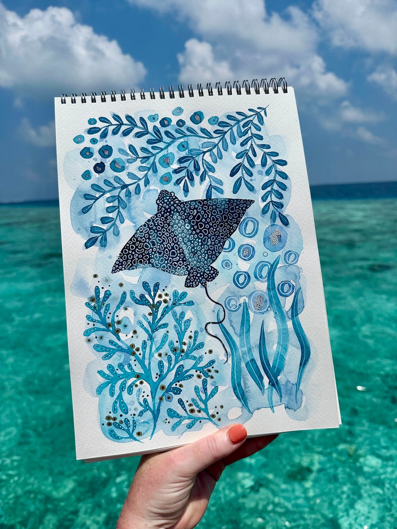 May include: Watercolor painting of a blue stingray with white spots, surrounded by blue and white seaweed and bubbles. The painting is on a white sheet of paper with a spiral binding.