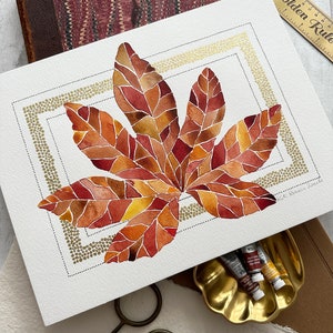 May include: A watercolor painting of a large, red, orange, and yellow leaf with a gold dot border. The leaf is framed by a double border, one in gold and one in black.