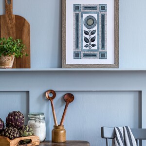 May include: A framed print of a blue and white stylized flower with geometric patterns on a light blue wall above a wooden table with a book, wooden spoons, a jar of flour, and artichokes.