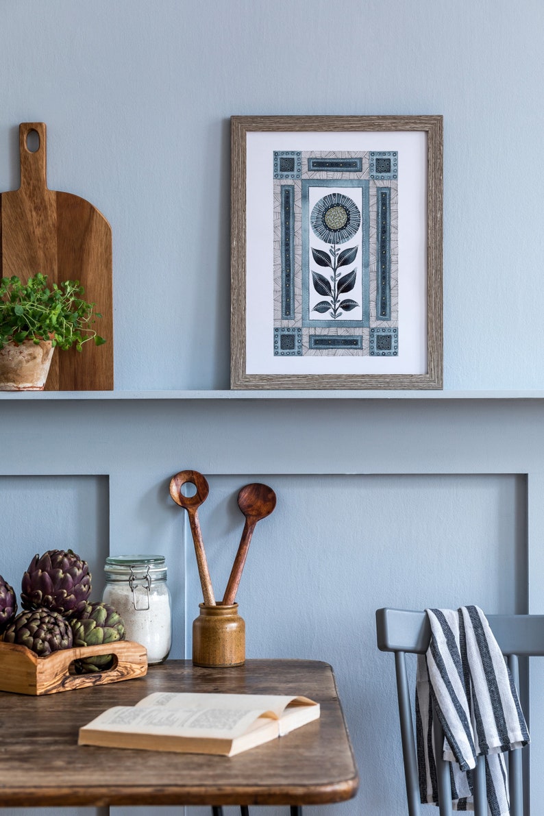 May include: A framed print of a blue and white stylized flower with geometric patterns on a light blue wall above a wooden table with a book, wooden spoons, a jar of flour, and artichokes.
