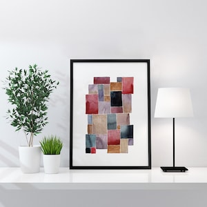 May include: A framed abstract watercolour art print with a collage of overlapping squares in shades of red, brown, black, and grey. The artwork is displayed on a white shelf with a small potted plant and a black desk lamp.