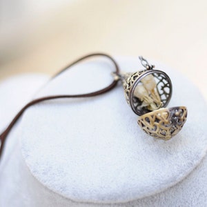 May include: A gold-toned pendant necklace with a delicate, openwork design. The pendant is shaped like a small cage and contains dried flowers. The necklace is made of brown leather cord.