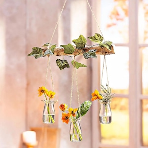 Könnte beinhalten: Ein rustikaler Holzast mit drei Glasflaschen, die daran hängen. Jede Flasche ist mit gelben Blumen und Grün gefüllt. Der Ast ist mit grünem Efeu verziert.