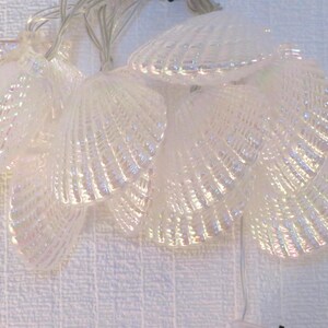 Op de afbeelding: Een reeks iriserende feeërieke lichtjes in de vorm van schelpen. De lichtjes zijn wit met een parelmoerglans, die op echte schelpen lijken. De draad is dun en zilverkleurig, en de lichtjes zijn dicht bij elkaar. Ideaal voor kust- of strandthema's.