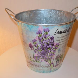 Op de afbeelding: Een gegalvaniseerde metalen emmer met twee gebogen handvatten. De emmer is versierd met een lavendelontwerp, met paarse bloemen en de woorden "Lavande de Provence".
