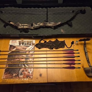 May include: A compound bow with a camouflage pattern resting in a black hard case. Included are six gold-colored arrows with purple fletching, a black quiver, a Delta Tru-Life instruction manual, and a small black accessory component.