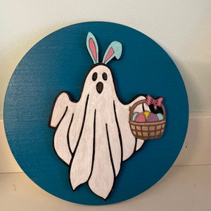 May include: A white ghost with blue bunny ears holding a brown basket filled with colourful Easter eggs on a teal blue background.