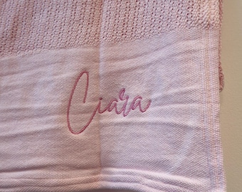May include: A light pink knitted blanket featuring a textured weave pattern and a smooth border. The name Ciara is embroidered in a darker pink cursive script on the lower section of the fabric, creating a personalized detail on the textile.