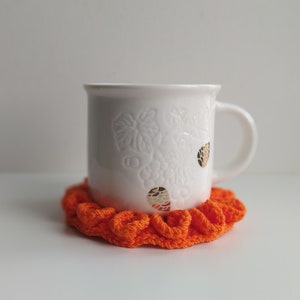 May include: A white ceramic mug with a handle, featuring a grape vine design and gold accents, sits on a bright orange, ruffled crochet coaster. The mug and coaster are on a white surface.