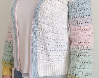 Puede incluir: Un cárdigan blanco de ganchillo con mangas de colores pastel. Las mangas hacen la transición de azul claro a rosa, luego a verde y terminan con un puño amarillo. El cárdigan tiene un ribete azul claro y rosa.