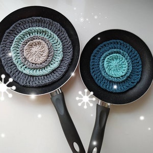 May include: Two black frying pans with crocheted pot holders in shades of blue and gray. The pot holders are circular and have a multi-layered design.