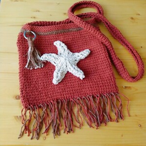 May include: A handmade, terracotta-coloured crochet bag with a long strap. The bag features a white starfish applique and a fringe along the bottom. A small tassel hangs from a metal ring near the top. The bag is made of a textured yarn.