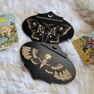 May include: Two black wooden ornaments shaped like moths with gold accents. The moths are depicted with intricate details and a crescent moon design. The ornaments are on a white fluffy surface.