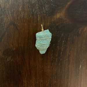 May include: A teal blue gemstone pendant wrapped in gold wire. The pendant is on a brown wooden surface.