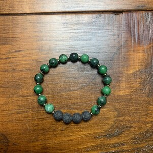 May include: A green and black beaded bracelet with gold accents. The bracelet is made with green gemstone beads and black lava rock beads.