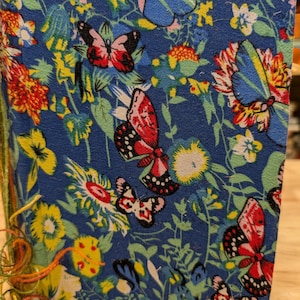 May include: A blue journal with a floral and butterfly pattern. The cover features red, white, and blue butterflies, yellow and orange flowers, and green leaves. The binding is secured with colorful thread.
