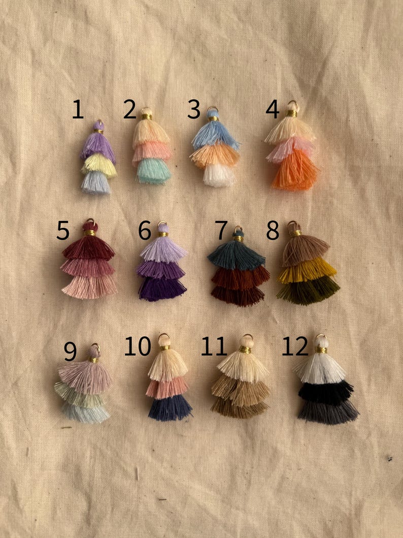 May include: Twelve different colored tassel charms with gold tops. The tassels are layered in three tiers and are arranged in rows of four.
