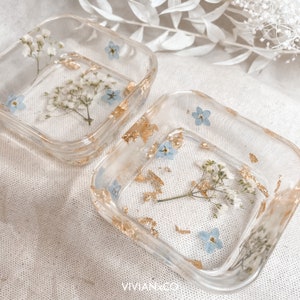 May include: Two square clear resin trinket dishes with gold leaf and pressed flowers. The dishes feature small blue flowers and white flowers.