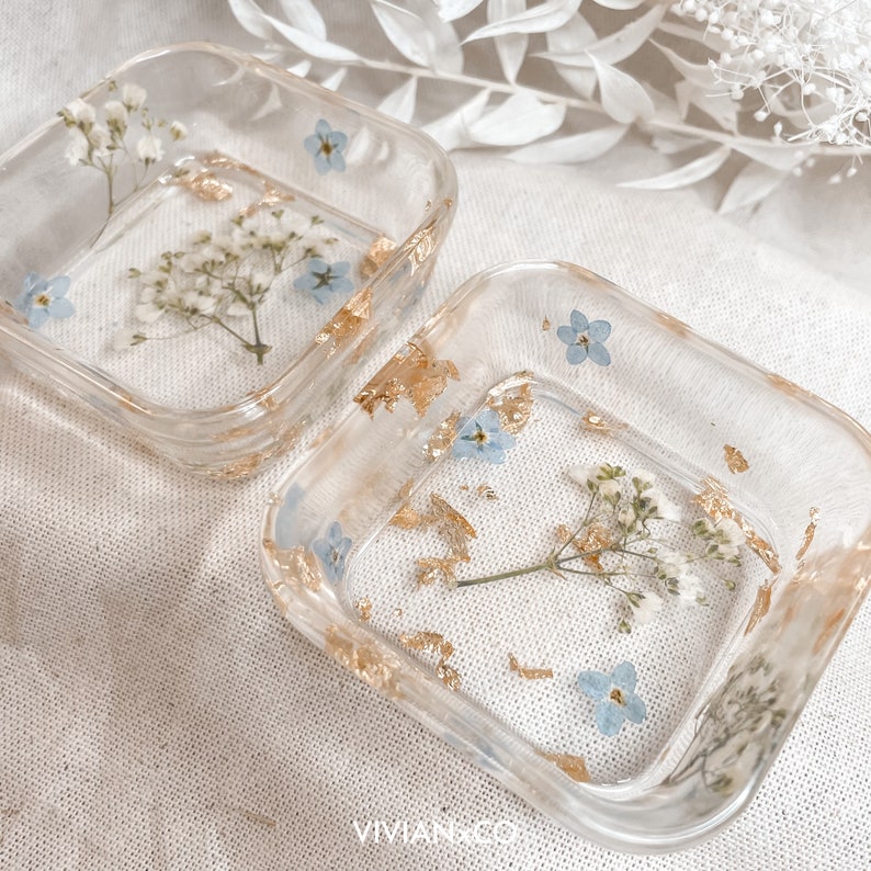 May include: Two square clear resin trinket dishes with gold leaf and pressed flowers. The dishes feature small blue flowers and white flowers.