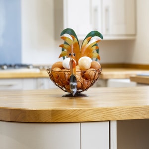 May include: A rooster-shaped wire egg holder with a brown, orange, and green design holds a collection of brown and white eggs on a wooden countertop.