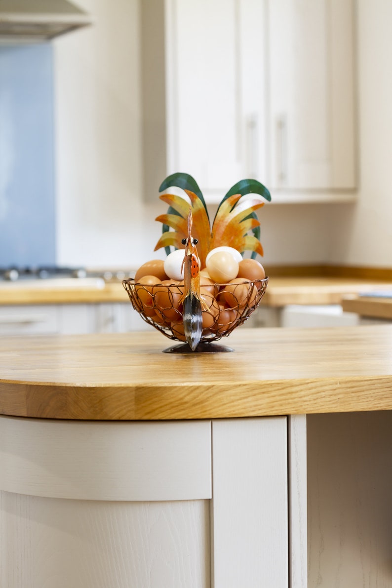 May include: A rooster-shaped wire egg holder with a brown, orange, and green design holds a collection of brown and white eggs on a wooden countertop.