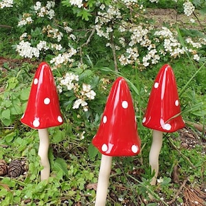 May include: Three red and white ceramic mushrooms with white polka dots. The mushrooms have a white stem and are arranged on a bed of green leaves and brown mulch.