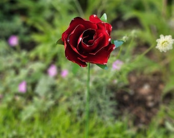 May include: A red metal rose with a green stem, standing in a grassy area with other flowers.