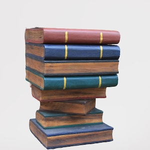 May include: A pile of decorative books in shades of red, blue, and green. Each book features gold detailing and a distressed look, giving them an antique or vintage feel. The books are stacked in a somewhat unstable arrangement.