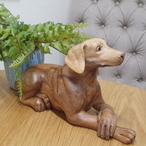 May include: A hand-carved wooden dog sculpture in a relaxed pose. The dog is a light brown colour, with lighter ears and snout. The sculpture is detailed, with visible paws and a relaxed posture. The dog is lying on a wooden surface.