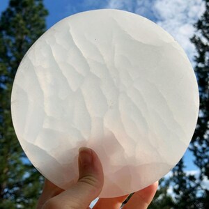 Selenite charging plate held to the light outdoors with blue sky, white clouds, and tree silhouettes