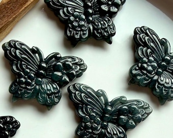 May include: Dark green butterfly-shaped carvings with intricate details, arranged on a white surface. The carvings have a polished finish and are likely made of stone or a similar material. A wooden block and a white crystal are in the background.