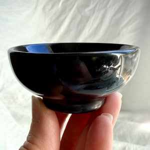May include: A small, smooth, solid black bowl with a rounded shape and a circular base. The bowl features a glossy, reflective surface that catches light, creating bright highlights along the rim and the curved exterior of the vessel.