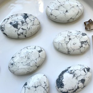 May include: A collection of white Howlite stones with dark gray veining, arranged on a white surface. The stones are oval-shaped and vary in size. Additional crystals, including a star-shaped one, are also present.