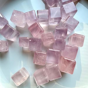 May include: A collection of polished, light purple and pink crystal cubes arranged on a white, shallow dish. The crystals vary in shade, with some appearing more translucent than others. A small, blue, teardrop-shaped stone and clear quartz crystals are also present.