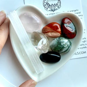 May include: A heart-shaped white ceramic dish filled with various polished crystals, including rose quartz, jasper, and clear quartz. A white selenite wand and a card with text that reads "WILD MOUNTAIN CRYSTALS" are also present.