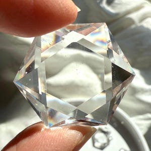 May include: A clear, faceted, hexagonal crystal with rainbow iridescence. The crystal is held in a hand.