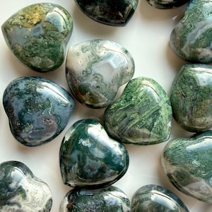 May include: A collection of heart-shaped moss agate stones. The stones display a mix of green, white, and clear colors, with unique patterns. The polished surfaces reflect light, highlighting the natural beauty of the gemstones. The arrangement suggests a focus on natural crystals and home decor.