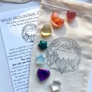May include: A small white pouch with a drawstring closure containing a variety of crystals. The pouch is on a white surface. The crystals are in various shapes and colors, including a green star, a purple heart, a clear star, a yellow heart, and a red and orange crescent moon. The pouch has a black and white illustration of a mountain range with a sun and moon.